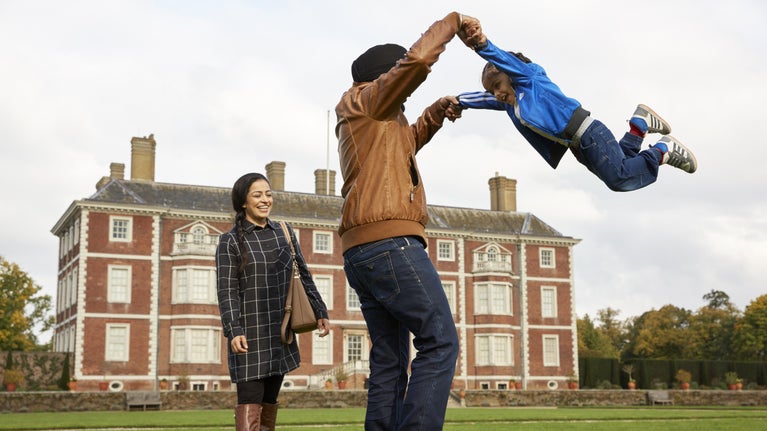 Father swinging daughter by the arms on lawn in front of Ham House while mother looks on